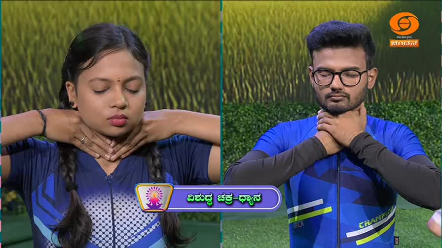 A young woman with braided hair and a man wearing glasses hold their hands to their throats, eyes closed. They are both dressed in blue athletic wear, likely participating in a yoga session broadcast on DD Chandana.