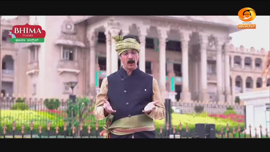 A man in traditional Indian attire stands before a grand building, gesturing with his hands. The DD Chandana logo is visible in the corner.