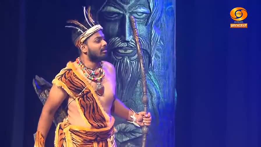 A performer in a tiger-striped costume and feathered headdress holds a staff, standing before a large, carved wooden figure. The DD Chandana logo appears in the corner, indicating a broadcast from India.
