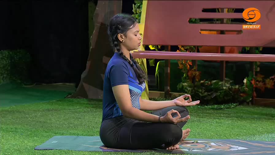 A young woman sits in meditation on a yoga mat, her eyes closed and fingers forming mudras. The DD Chandana logo is visible on a bench behind her, suggesting this is part of a broadcast from India.