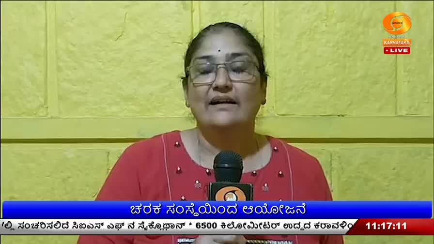 A woman speaks into a microphone, her red top adorned with small white dots. A graphic overlay at the bottom of the screen displays text in Kannada and the time.