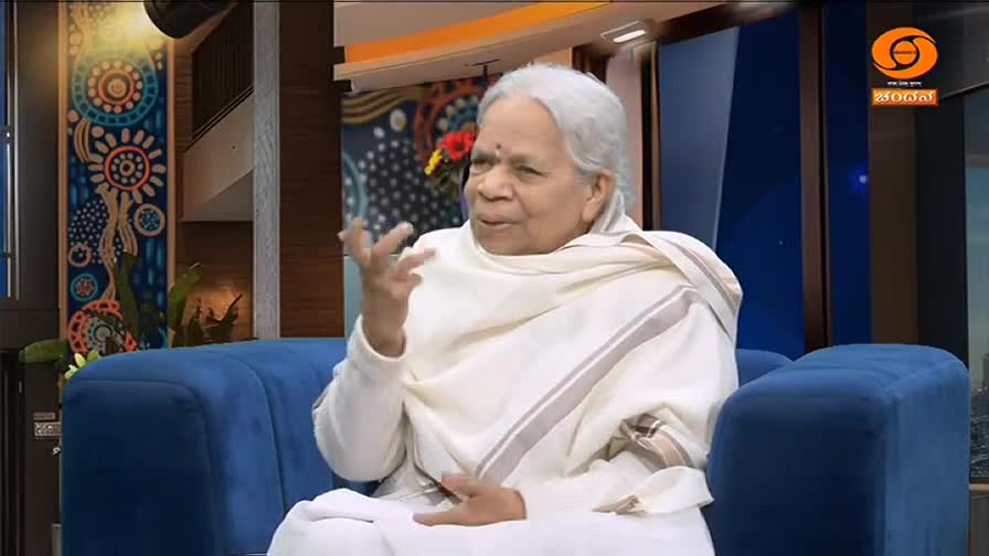 An older woman, dressed in white, speaks animatedly on a blue couch. The DD Chandana logo is visible in the background.
