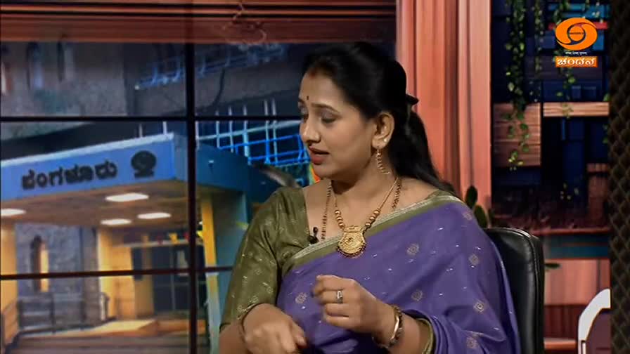 A woman in a purple sari speaks animatedly, her hands gesturing. Behind her, a backdrop displays the DD Chandana logo and text in Kannada, hinting at a broadcast from India.