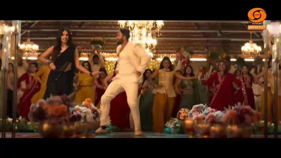 A man in a white kurta moves with energy, his arms outstretched. Beside him, a woman in a black sari mirrors his dance, her expression bright. A crowd of dancers in vibrant Indian attire sways behind them.