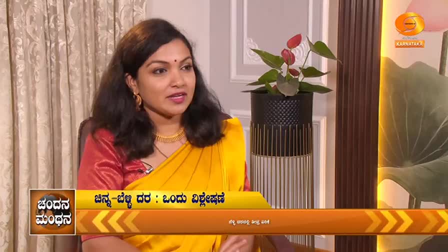 A woman in a yellow sari and red blouse speaks, her gold necklace catching the light. Behind her, a potted plant with red flowers sits on a patterned stand.
