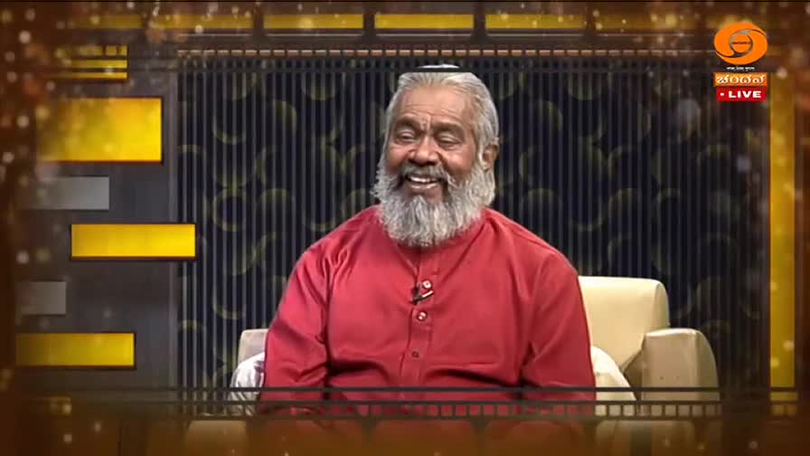 A man with a long white beard, wearing a bright red shirt, laughs heartily on a television set. The DD Chandana logo and "LIVE" indicator are visible in the corner.