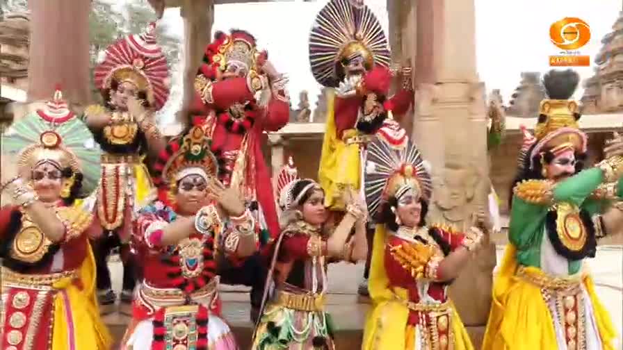 A group of performers in elaborate costumes and headdresses move in unison. Their hands are raised, and their bodies are turned towards the viewer, as if presenting a scene from DD Chandana.