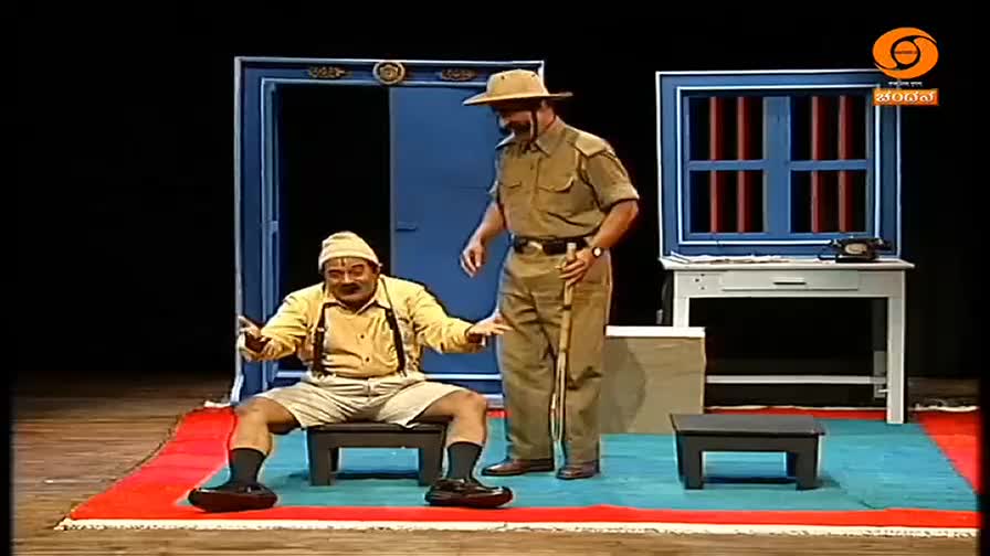 A man in a yellow shirt and shorts sits on a stool, gesturing with his hands. A uniformed man with a hat stands over him, holding a cane. This looks like a scene from a DD Chandana production.