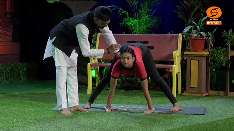A man in a white kurta and dark vest guides a young girl in a yoga pose on a mat. The DD Chandana logo is visible in the background.