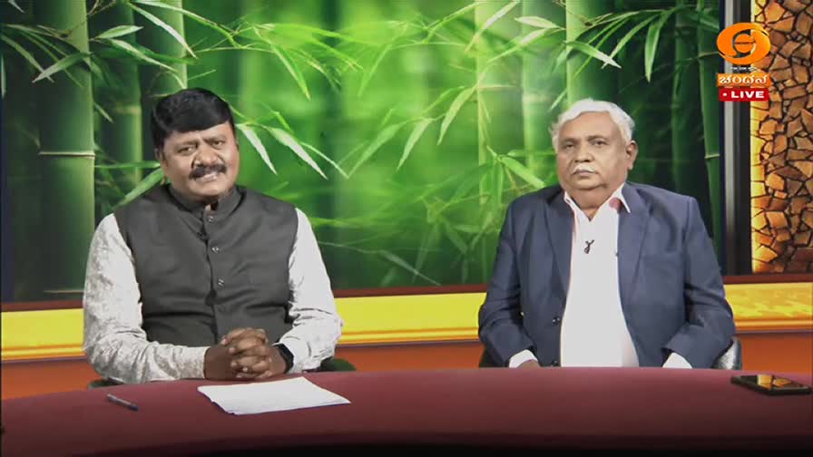 Two men sit at a table in front of a green, bamboo-themed backdrop. The man on the left, wearing a dark vest over a white shirt, gestures with his hands clasped. The man on the right, in a blue suit and white shirt, looks directly ahead.