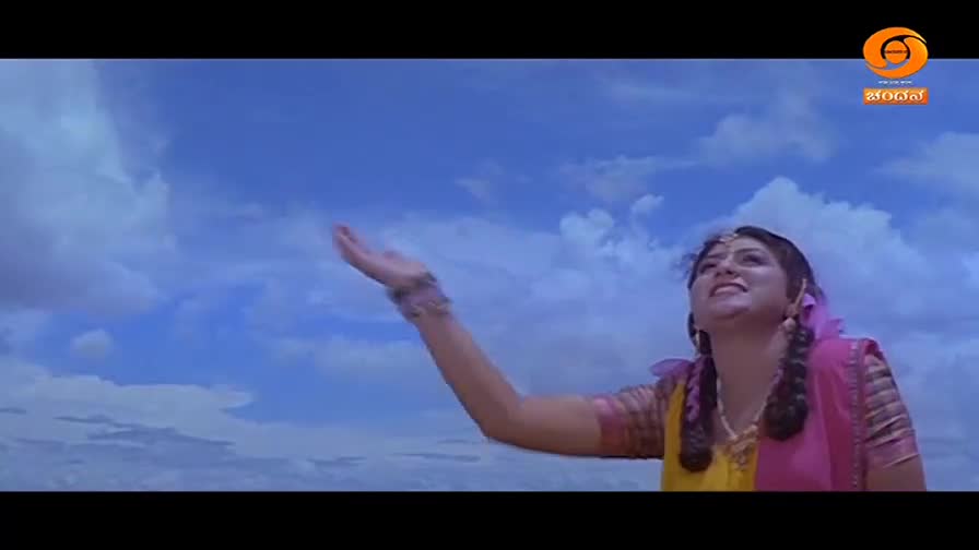 A young woman in a vibrant yellow and pink sari reaches her hand up towards the vast blue sky. Her expression is one of hopeful anticipation as she looks upward. The DD Chandana logo is visible in the corner.