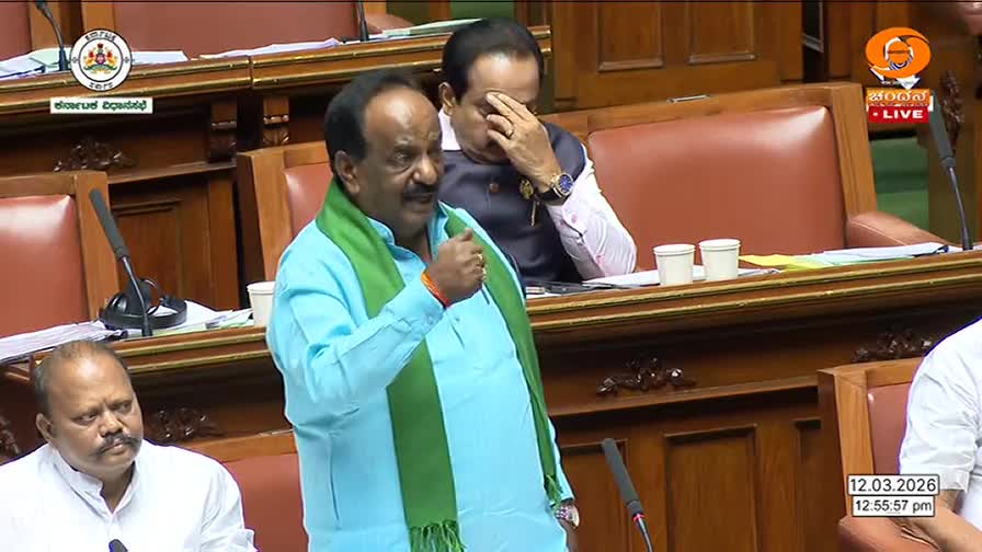 A man in a light blue kurta and green sash gestures emphatically while speaking. Behind him, another man rubs his forehead, looking weary.