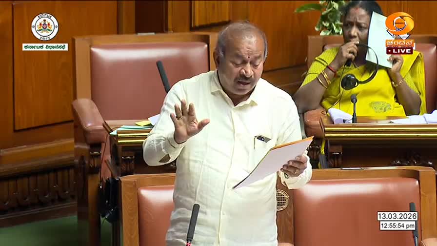 A man in a light-colored shirt speaks, holding a document. Behind him, a woman in a bright yellow sari gestures with a paper.