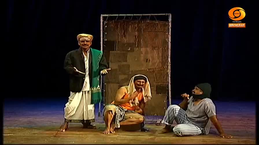 A man in a white dhoti and green shawl stands center stage, gesturing with his hands. Two other men, one in an orange vest and the other in a grey shirt, are seated on the floor before him. The DD Chandana logo is visible in the corner.