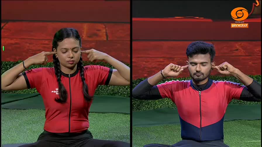 A woman and a man, both in red and black athletic wear, sit cross-legged and place their index fingers over their ears. This demonstration, likely part of a yoga segment on DD Chandana, focuses on a moment of quiet contemplation.