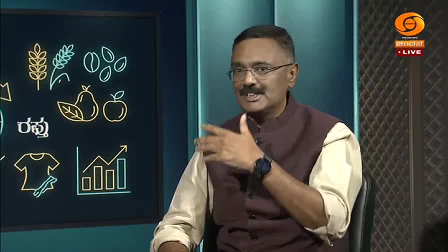 A man in a maroon vest over a cream shirt gestures with his right hand, speaking animatedly. Behind him, a screen displays neon graphics of agricultural products and a rising bar chart, with the DD Chandana logo and "LIVE" visible.