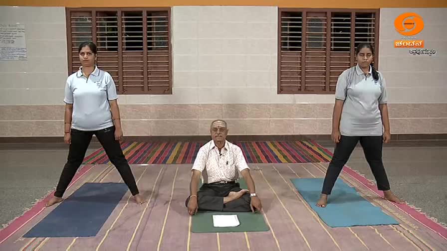 Two women stand with their legs spread wide on yoga mats, mirroring each other's posture. A man sits cross-legged between them, guiding the session for DD Chandana.