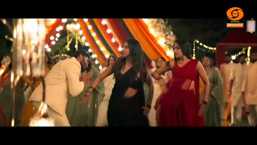 A man in a cream suit bows to a woman in a black sari as she dances. Beside her, another woman in a red sari moves to the music, surrounded by guests.