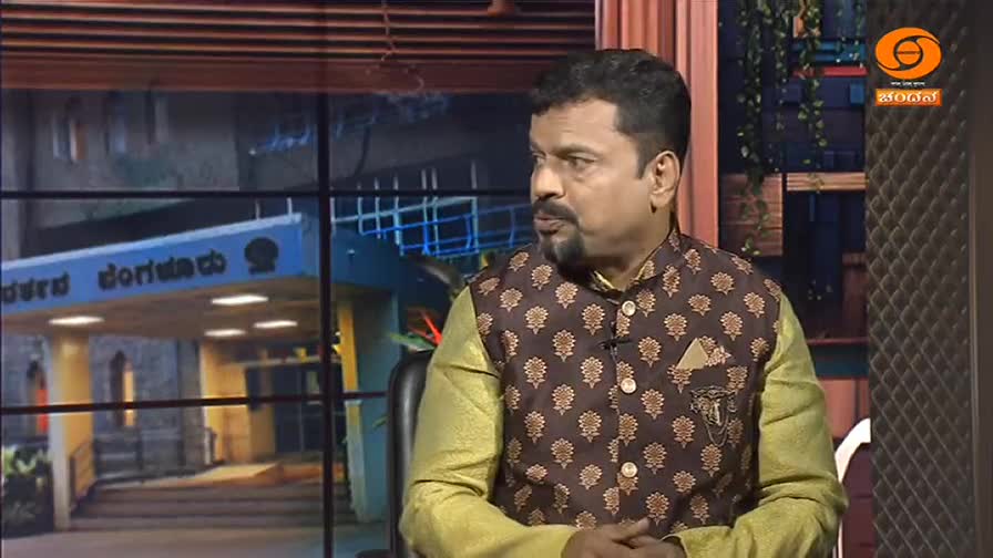 A man in a patterned vest speaks on a television program, the DD Chandana logo visible in the corner. Behind him, a building facade with Kannada script is displayed on a screen.