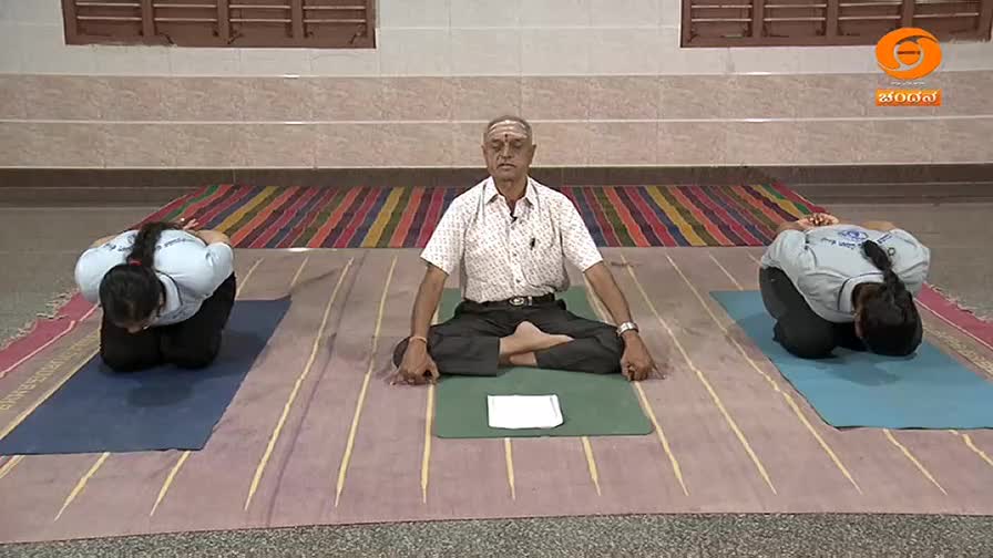 A man in a patterned shirt sits cross-legged, eyes closed, while two young women in white shirts and black pants kneel with their heads bowed. This yoga session is being broadcast on DD Chandana. A man in a patterned shirt sits cross-legged, eyes closed, while two young women in white shirts and black pants kneel with their heads bowed. This yoga session is being broadcast on DD Chandana.
