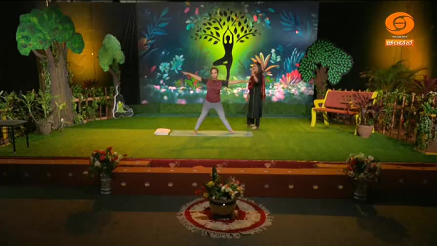 A woman in a maroon top and grey leggings holds a wide-legged yoga pose on a mat. Another woman stands beside her, speaking into a microphone.