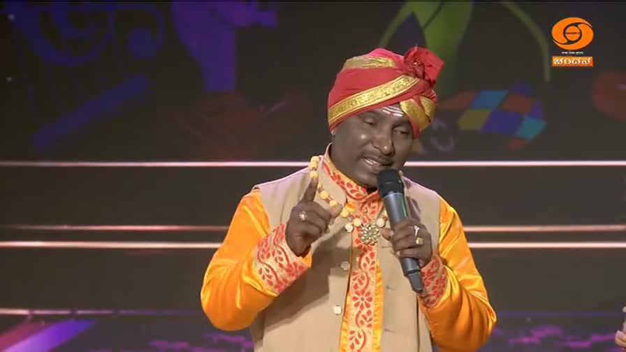 A man in a vibrant yellow kurta and red turban speaks into a microphone, gesturing with his right hand. He's on a stage, likely for a DD Chandana broadcast in India.