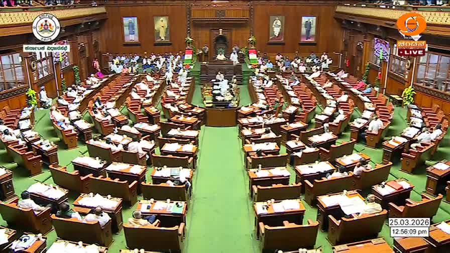 The assembly hall is packed with people seated in rows of wooden desks. A man stands at a podium in the center, addressing the gathered members.