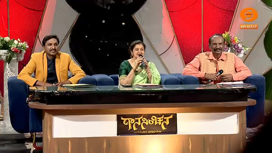 Three people sit on a blue couch behind a wooden desk. The woman in the center, wearing a green sari, holds a microphone and speaks. To her left, a man in a yellow jacket looks on, and to her right, a man in an orange shirt holds a microphone. A sign with Kannada script sits on the desk in front of them.