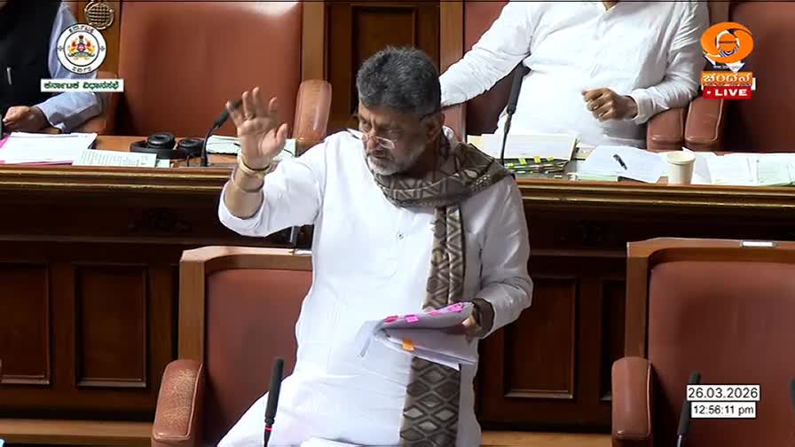 A man in a white kurta and a patterned scarf raises his hand, holding papers. He stands before a wooden desk in a room filled with similar seating, likely a legislative assembly.