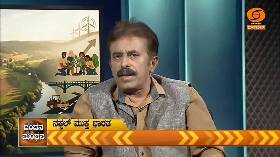 A man with a mustache speaks on DD Chandana, his backdrop displaying an illustrated landscape. The graphic shows wind turbines, a bridge, a road roller, and people planting a tree, all under an upward-trending arrow.