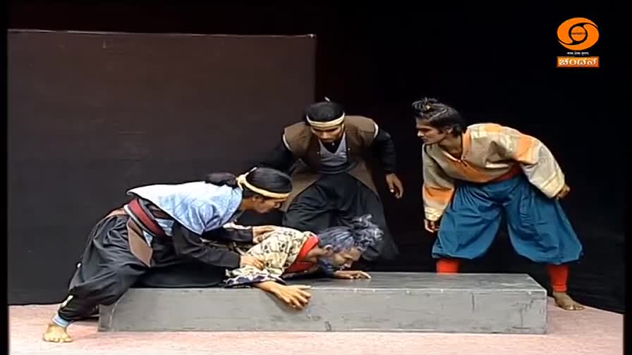 Two men in traditional Indian attire are helping a third man, who is on his hands and knees. Another man stands nearby, watching intently.