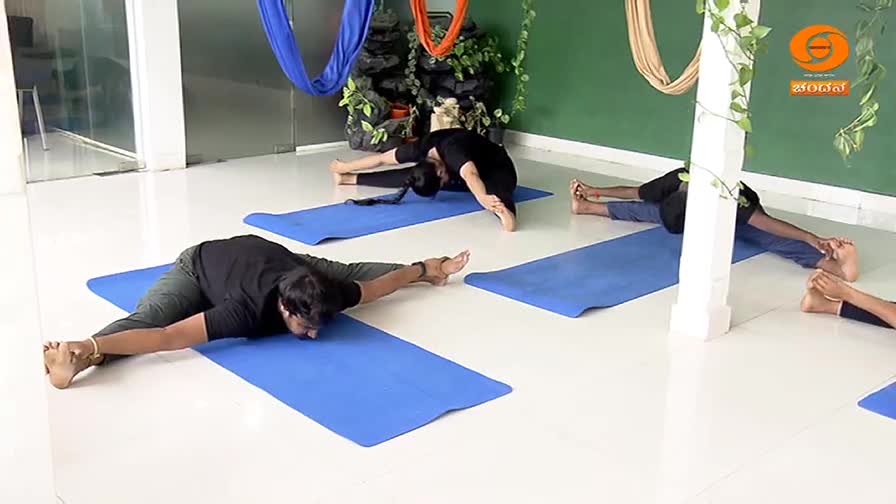 Several people are holding yoga poses on blue mats. The man in the foreground stretches his legs wide, reaching forward with his torso. On DD Chandana, a yoga instructor guides them through the practice.