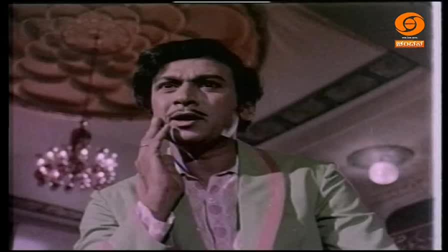 A man with a mustache, dressed in a light suit and patterned shirt, touches his cheek with his fingers. The DD Chandana logo appears in the corner, indicating this is from an Indian broadcast.