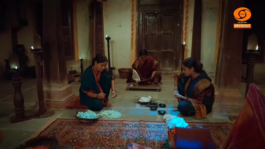 Three women are gathered on the floor, sorting through piles of white jasmine flowers. The warm glow of oil lamps illuminates the scene, casting long shadows across the patterned rug.