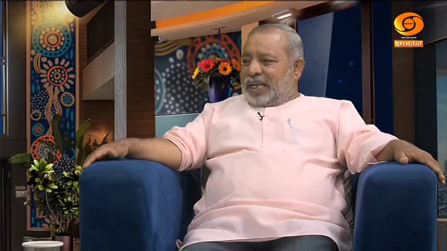 A man in a pale pink kurta sits in a blue armchair, speaking. Behind him, a colorful mural adorns the wall, and a vase of flowers sits on a shelf. The DD Chandana logo is visible in the corner of the screen. A man in a pale pink kurta sits in a blue armchair, speaking. Behind him, a colorful mural adorns the wall, and a vase of flowers sits on a shelf. The DD Chandana logo is visible in the corner of the screen.