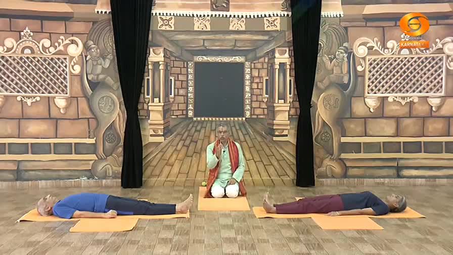 A man in a light green kurta and saffron shawl sits cross-legged on an orange mat, gesturing with his hands. Two individuals lie flat on their backs on similar mats, one in a blue t-shirt and the other in a dark t-shirt and maroon pants. The backdrop is a painted mural of an ancient Indian hall. A man in a light green kurta and saffron shawl sits cross-legged on an orange mat, gesturing with his hands. Two individuals lie flat on their backs on similar mats, one in a blue t-shirt and the other in a dark t-shirt and maroon pants. The backdrop is a painted mural of an ancient Indian hall.