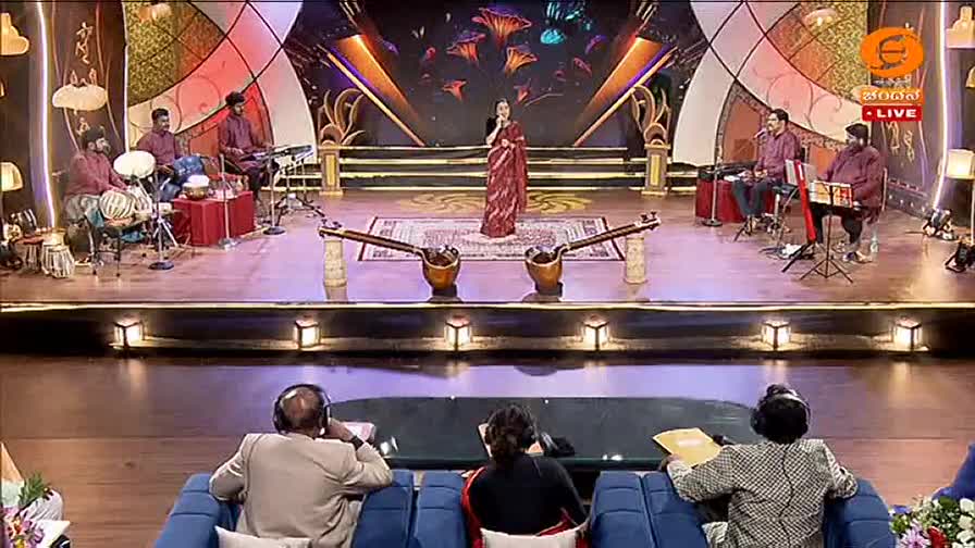 A woman in a red sari stands center stage, singing into a microphone. Musicians behind her play traditional Indian instruments, their rhythmic beats filling the air. Judges sit in the foreground, listening intently. A woman in a red sari stands center stage, singing into a microphone. Musicians behind her play traditional Indian instruments, their rhythmic beats filling the air. Judges sit in the foreground, listening intently.