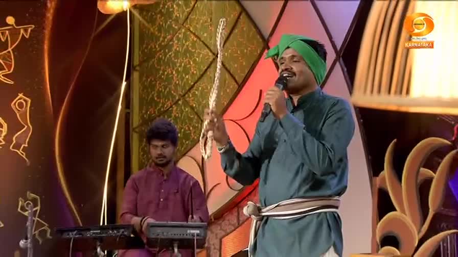A man in a green turban sings into a microphone, holding a beaded string. Behind him, another man plays a keyboard. A man in a green turban sings into a microphone, holding a beaded string. Behind him, another man plays a keyboard.