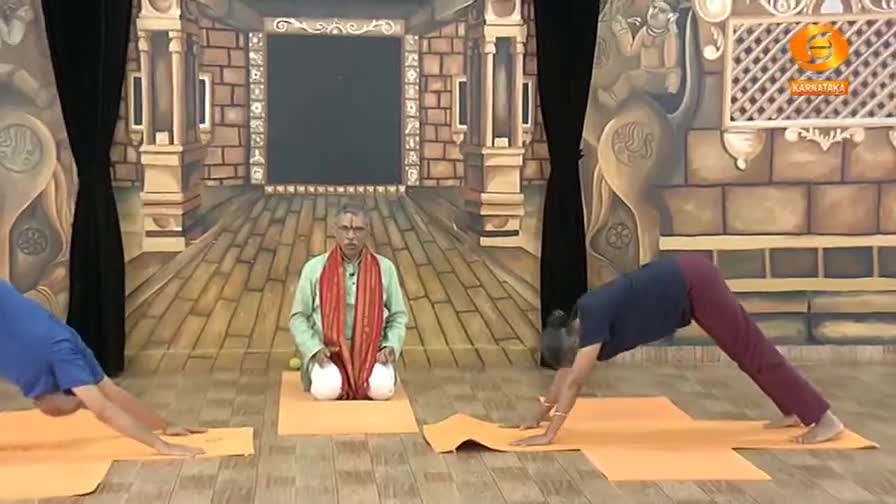 Two people are on orange mats, holding a downward-facing dog pose. A man in traditional Indian attire sits cross-legged between them, observing. Two people are on orange mats, holding a downward-facing dog pose. A man in traditional Indian attire sits cross-legged between them, observing.