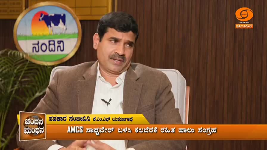 A man in a brown jacket speaks on a television program, the DD Chandana logo visible in the corner. A circular emblem with a cow and the word "Nandini" hangs behind him. A man in a brown jacket speaks on a television program, the DD Chandana logo visible in the corner. A circular emblem with a cow and the word "Nandini" hangs behind him.