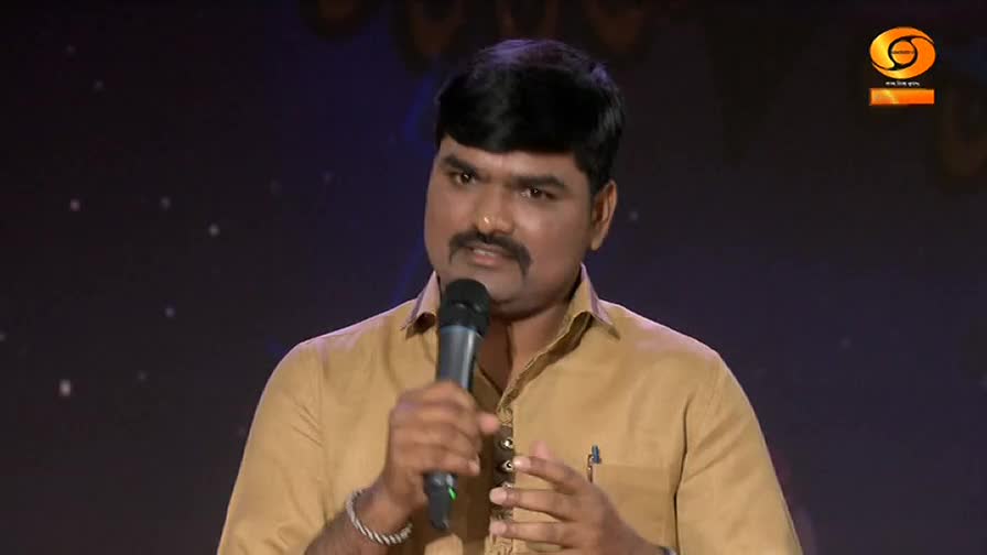 A man in a tan shirt speaks into a microphone, his left hand gesturing. The DD Chandana logo appears in the upper right corner. A man in a tan shirt speaks into a microphone, his left hand gesturing. The DD Chandana logo appears in the upper right corner.