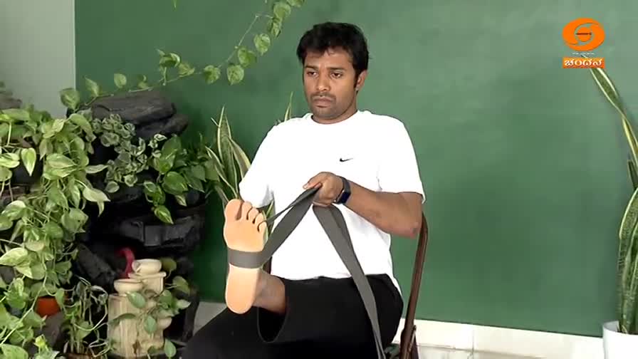 A man in a white t-shirt stretches his foot with a strap, his toes clearly visible. Lush green plants surround him, and a small logo for DD Chandana appears in the corner. A man in a white t-shirt stretches his foot with a strap, his toes clearly visible. Lush green plants surround him, and a small logo for DD Chandana appears in the corner.