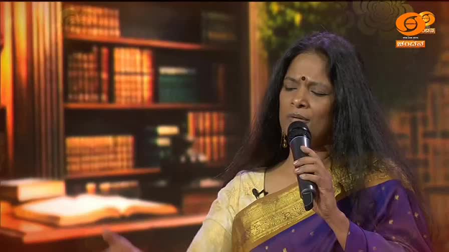 A woman in a vibrant purple sari sings into a microphone, her eyes closed in concentration. Behind her, bookshelves filled with volumes create a warm, inviting backdrop, hinting at a cultural broadcast from DD Chandana. A woman in a vibrant purple sari sings into a microphone, her eyes closed in concentration. Behind her, bookshelves filled with volumes create a warm, inviting backdrop, hinting at a cultural broadcast from DD Chandana.