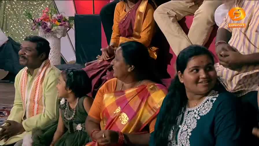A man in a light yellow kurta and a girl in a dark dress sit attentively. A woman in a vibrant orange and pink sari laughs beside them, while another woman in a dark blue top with white embroidery smiles. The DD Chandana logo is visible in the background. A man in a light yellow kurta and a girl in a dark dress sit attentively. A woman in a vibrant orange and pink sari laughs beside them, while another woman in a dark blue top with white embroidery smiles. The DD Chandana logo is visible in the background.