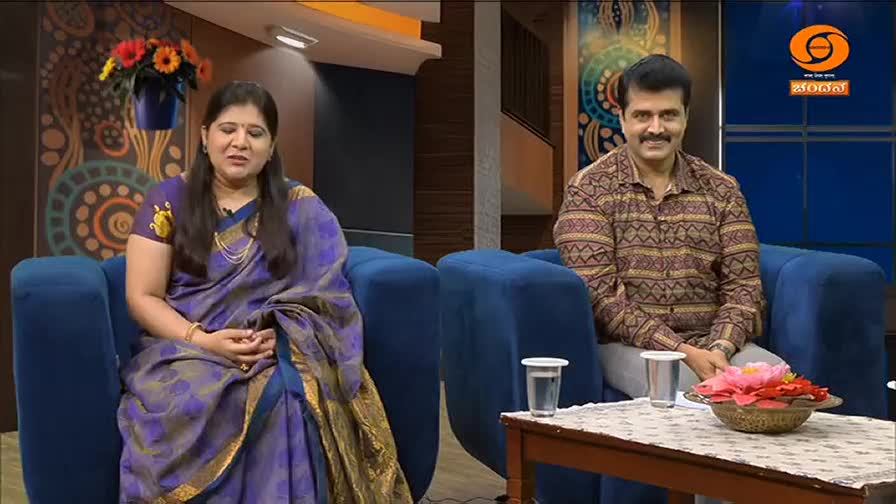 A woman in a purple silk saree and a man in a patterned shirt sit on blue chairs, facing forward. They are in a studio setting, with the DD Chandana logo visible in the background. A woman in a purple silk saree and a man in a patterned shirt sit on blue chairs, facing forward. They are in a studio setting, with the DD Chandana logo visible in the background.