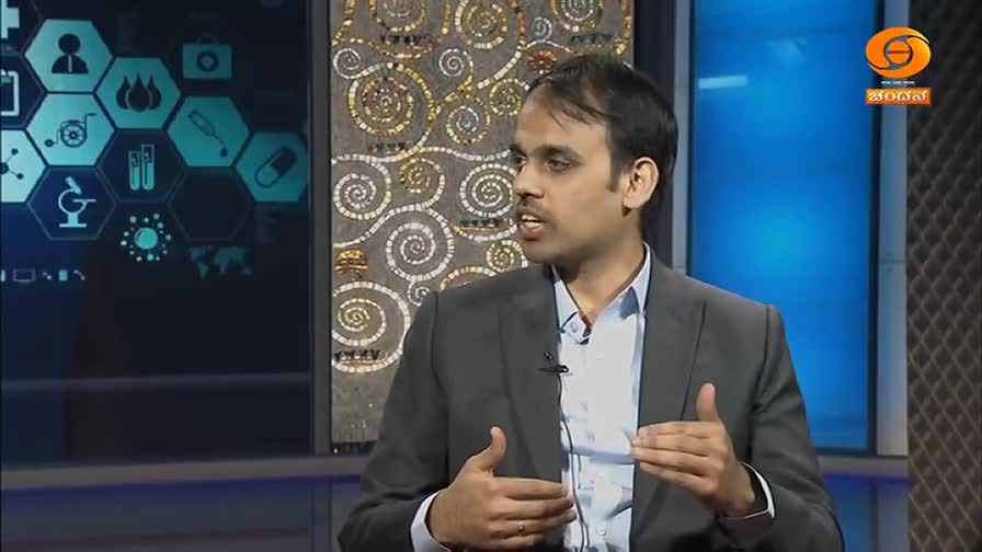 A man in a suit speaks animatedly, gesturing with his hands on a set with a DD Chandana logo. Behind him, a screen displays a grid of medical icons.