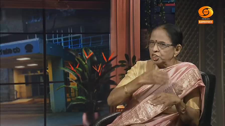 A woman in a pink sari speaks animatedly, her hands gesturing as she talks. Behind her, a window shows a building entrance at night, with a DD Chandana logo visible on a screen.