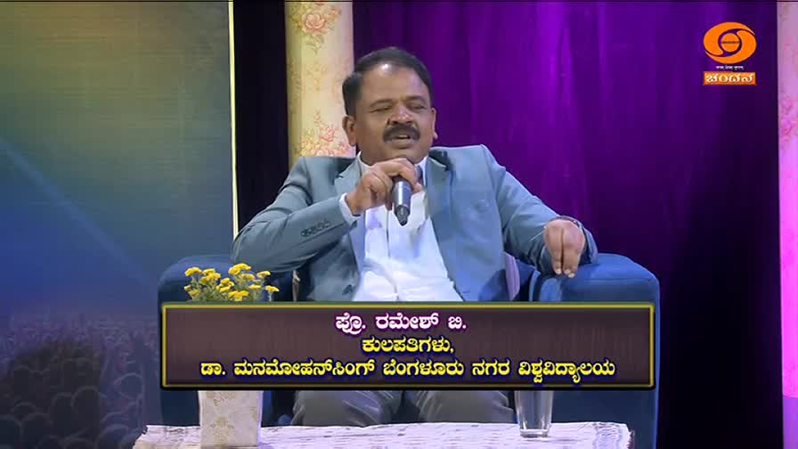 A man in a blue suit speaks into a microphone, seated on a dark blue chair. The DD Chandana logo appears in the upper right corner.