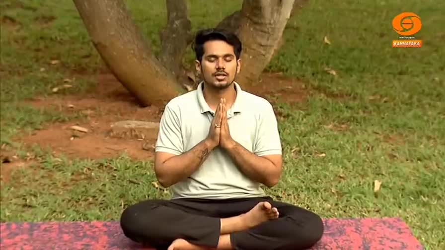 A man sits cross-legged on a mat in a park, his hands pressed together in prayer. He's on the grounds of Karnataka, likely for a DD Chandana broadcast.