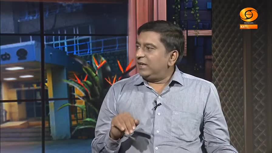 A man in a light blue shirt speaks animatedly, gesturing with his right hand. Behind him, a graphic of what looks like a building and vibrant orange and green plants fills the screen.